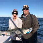 Woman smiling with mouth open holding a very large striped bass, captain beside her