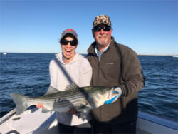 Woman smiling with mouth open holding a very large striped bass, captain beside her