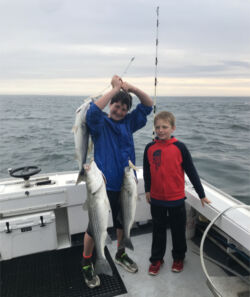 Two boys proudly holding three large striped bass on back deck of a boat