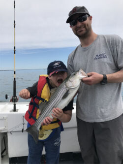 Boy and his dad holding striped bass with boy pretending to bite the fish