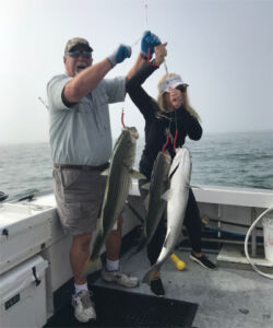 Woman holding up fishing rig with three large striped bass that she caught, with Captain Mike helping